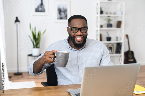 Smiling African-American businessman in glasses sitting at the desk, working from home, holding a cup of coffee and laughing, looking at the laptop screen, and participating in the webinar