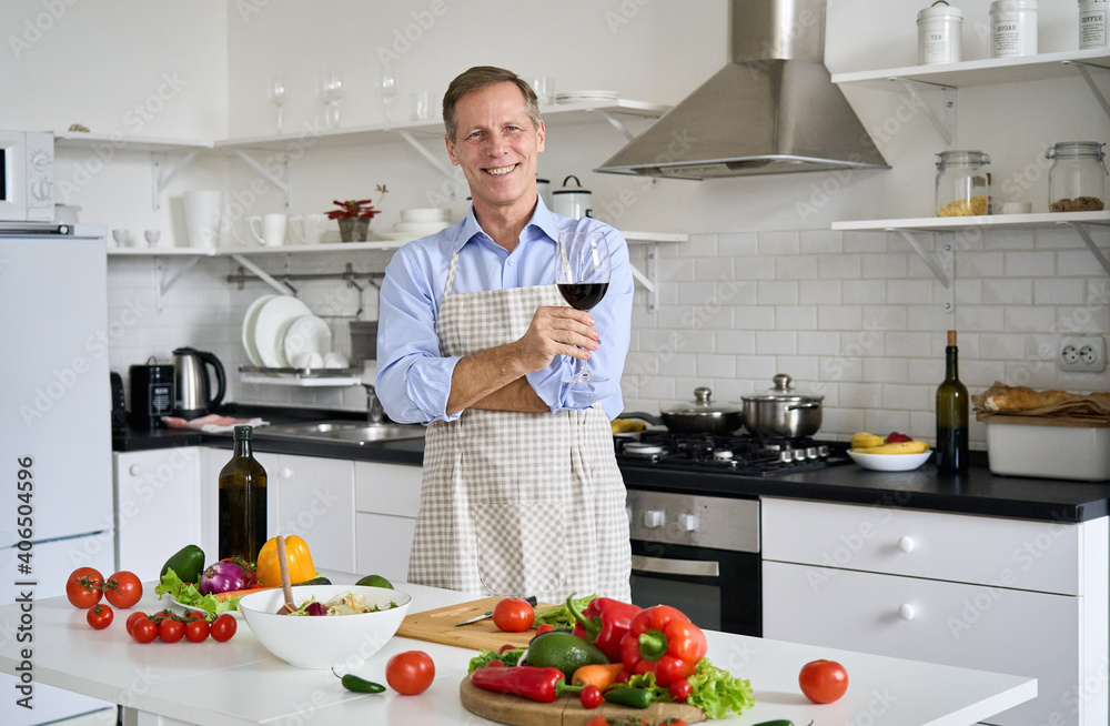 Happy senior old man holding glass of wine standing in kitchen enjoying cooking. Smiling middle aged 60s chef wearing apron looking at camera preparing healthy food vegetable salad at home, portrait. Happy senior old man holding glass of wine standing in kitchen enjoying cooking. Smiling middle aged 60s chef wearing apron looking at camera preparing healthy food vegetable salad at home, portrait.