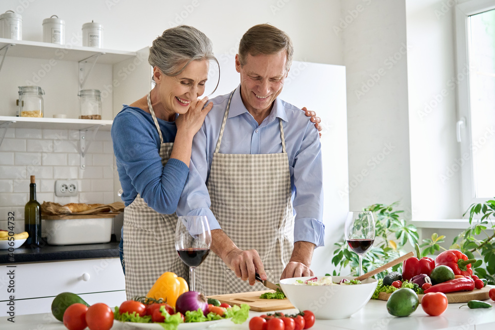 Happy old senior 50s couple wearing aprons having fun preparing meal salad at home. Smiling middle aged wife hugging older husband cutting fresh vegetables, cooking healthy food together in kitchen. Happy old senior 50s couple wearing aprons having fun preparing meal salad at home. Smiling middle aged wife hugging older husband cutting fresh vegetables, cooking healthy food together in kitchen.