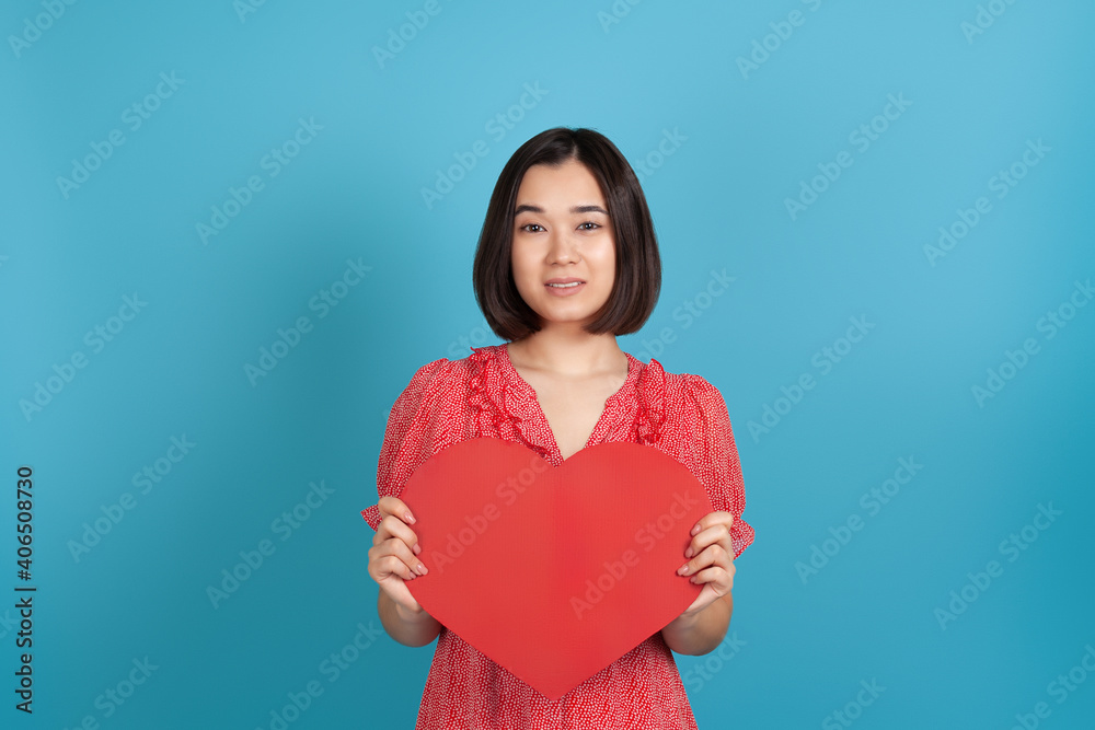 close-up smiling beautiful young Asian woman in a red dress holding a large red paper heart at her chest, isolated on a blue background. close-up smiling beautiful young Asian woman in a red dress holding a large red paper heart at her chest, isolated on a blue background.