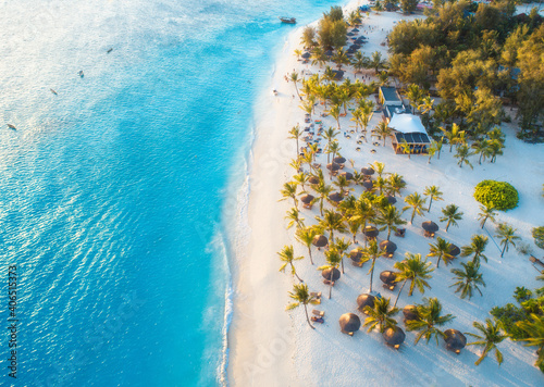 Aerial view of umbrellas, green palms on the sandy beach at sunset. Summer holiday in Zanzibar, Africa. Tropical landscape with palm trees, parasols, white sand, blue water, waves, people. Top view photo