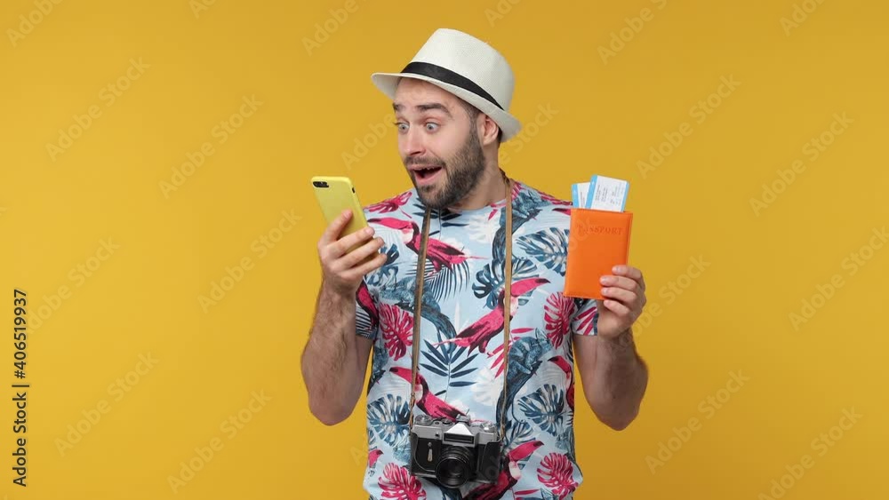 Excited happy traveler tourist young man in hat isolated on yellow background. Passenger travel on weekends. Air flight journey concept. Hold passport tickets using mobile cell phone book hotel taxi
