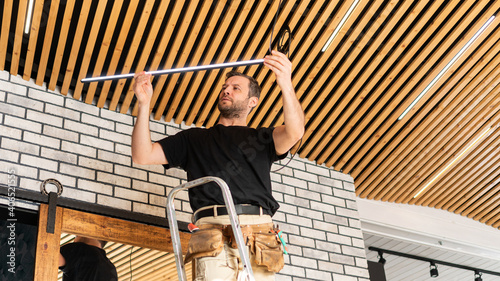 A male electrician changes lamps and repairs electrical wiring. A builder is installing a loft-style wooden plank ceiling. Installation of low-voltage LED lamps in the designer ceiling.