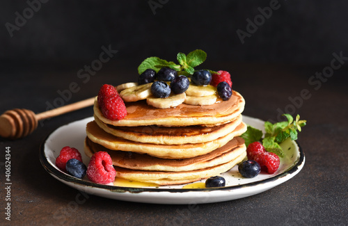 Homemade pancakes with banana, raspberries, blueberries and honey on a dark concrete background.