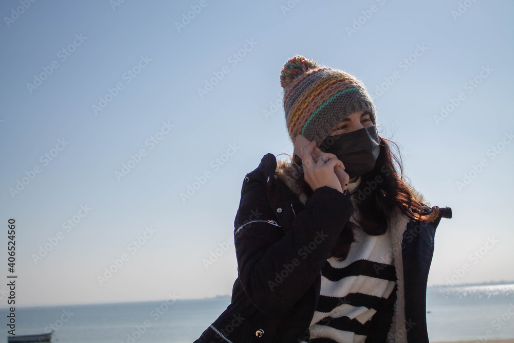 Beautiful girl with mask and hat next to the beach in winter talking on phone
