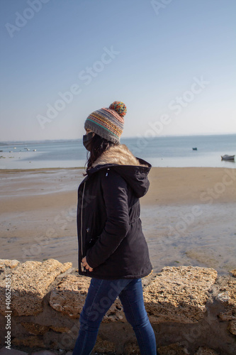 Beautiful girl with mask and hat next to the beach in winter with boats in the water 