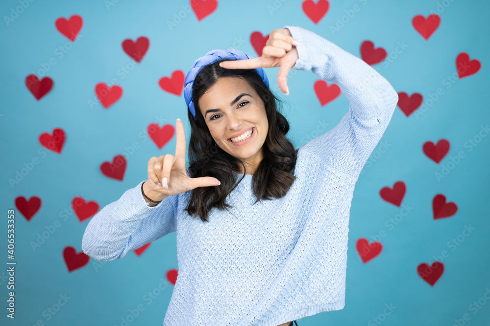 Young beautiful woman over blue background with red hearts smiling making frame with hands and fingers with happy face Young beautiful woman over blue background with red hearts smiling making frame with hands and fingers with happy face