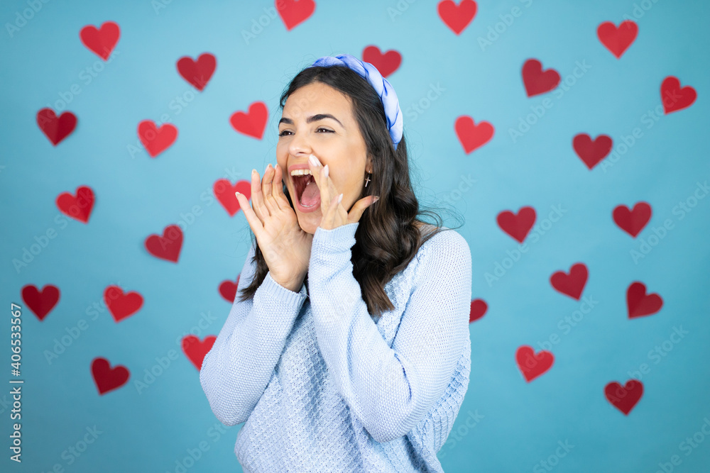 Young beautiful woman over blue background with red hearts shouting and screaming loud to side with hands on mouth Young beautiful woman over blue background with red hearts shouting and screaming loud to side with hands on mouth