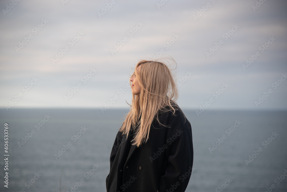 mujer rubia con abrigo negro mirando hacia el mar y tomando el aire en un día de invierno y cielo nublado mujer rubia con abrigo negro mirando hacia el mar y tomando el aire en un día de invierno y cielo nublado