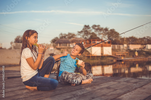Little boy and girl eating sandwiches and fishing on a lake, the boy showing gesture thumb up.