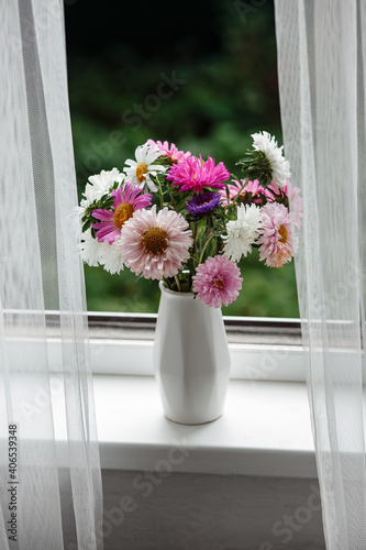 Purple and white flowers in a white vase on the windowsill behind the tulle.