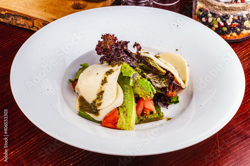 Italian caprese salad with sliced tomatoes, mozzarella cheese, pesto sauce, lettuce on white plate.