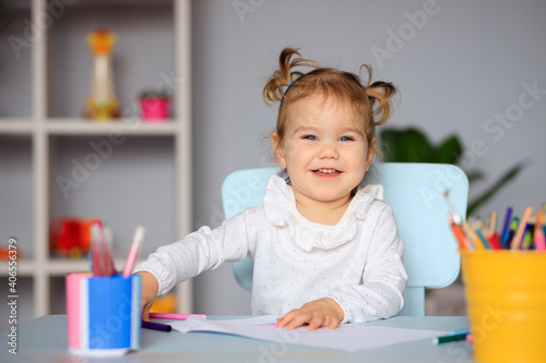 happy little girl sits at the table and draws on paper with colored pencils
