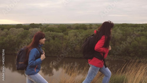 Female travelers with backpacks are walking along a high mountain and smiling. Girlfriends hiking trip. Life adventures of women in nature. Teamwork. Family weekend together by the flowing river