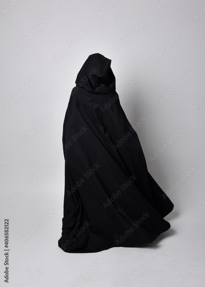 Full length portrait of pretty black haired woman wearing long dark gown nada cloak. Standing pose facing away from the camera, against a studio background. Full length portrait of pretty black haired woman wearing long dark gown nada cloak. Standing pose facing away from the camera, against a studio background.