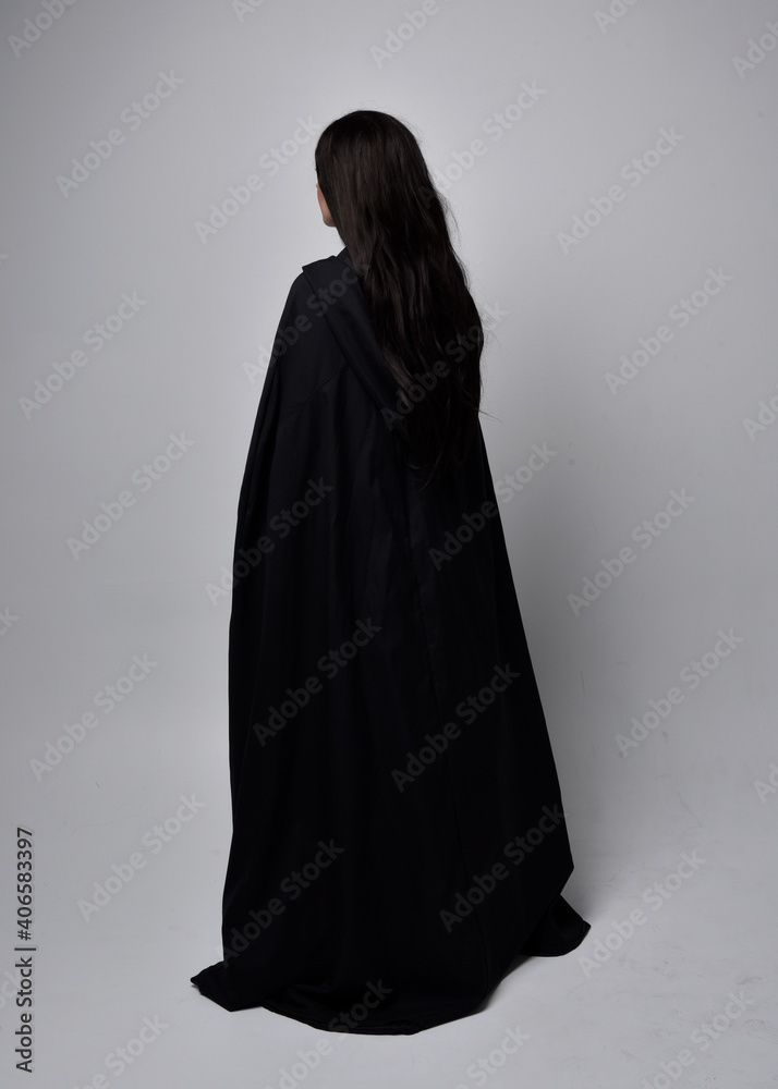 Full length portrait of pretty black haired woman wearing long dark gown nada cloak.  Standing pose facing away from the camera, against a  studio background.