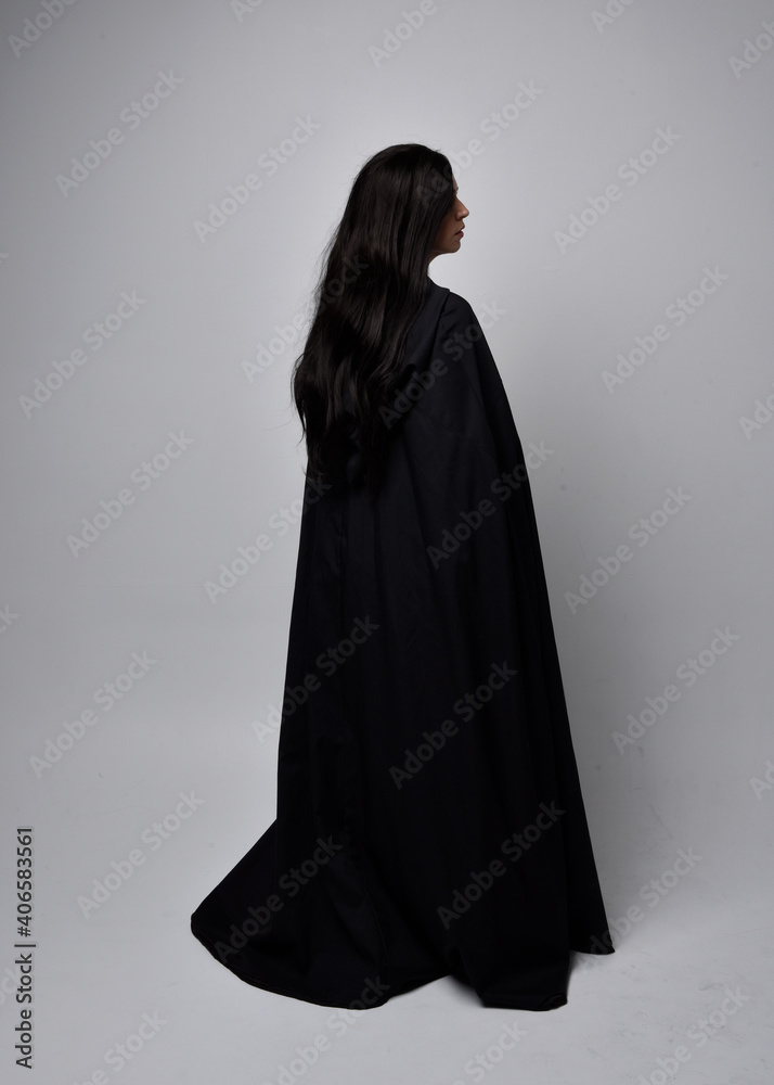 Full length portrait of pretty black haired woman wearing long dark gown nada cloak. Standing pose facing away from the camera, against a studio background. Full length portrait of pretty black haired woman wearing long dark gown nada cloak. Standing pose facing away from the camera, against a studio background.