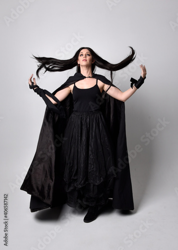 Full length portrait of pretty black haired woman wearing long dark gown and a cloak.  Standing pose facing away from the camera, against a  studio background.