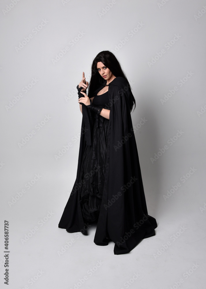 Full length portrait of pretty black haired woman wearing long dark gown and a cloak.  Standing pose facing away from the camera, against a  studio background.