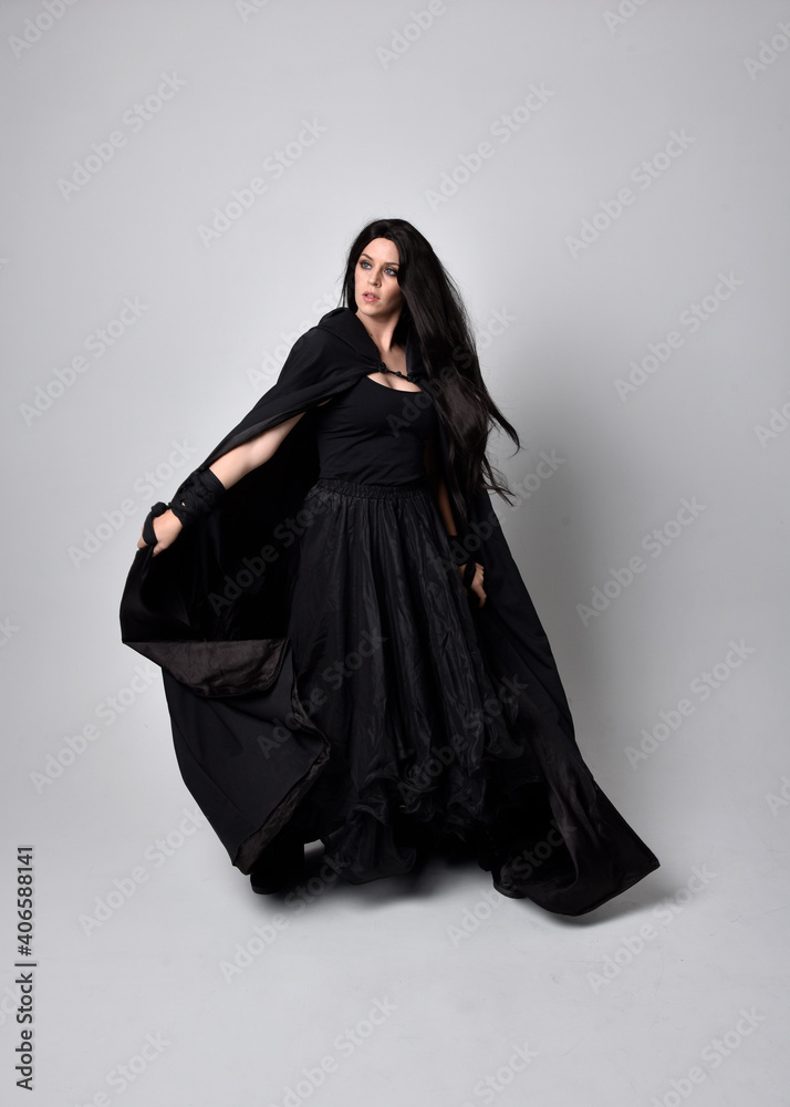 Full length portrait of pretty black haired woman wearing long dark gown and a cloak. Standing pose facing away from the camera, against a studio background. Full length portrait of pretty black haired woman wearing long dark gown and a cloak. Standing pose facing away from the camera, against a studio background.