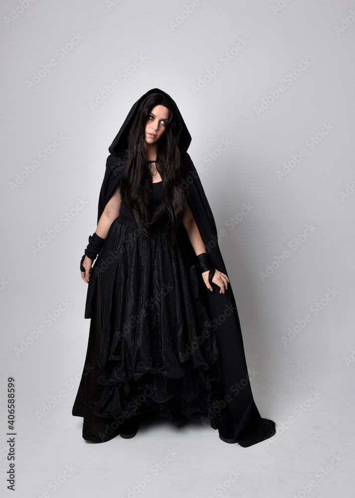 Full length portrait of pretty black haired woman wearing long dark gown and a cloak.  Standing pose facing away from the camera, against a  studio background.