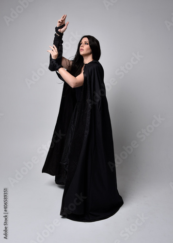 Full length portrait of pretty black haired woman wearing long dark gown and a cloak.  Standing pose facing away from the camera, against a  studio background.