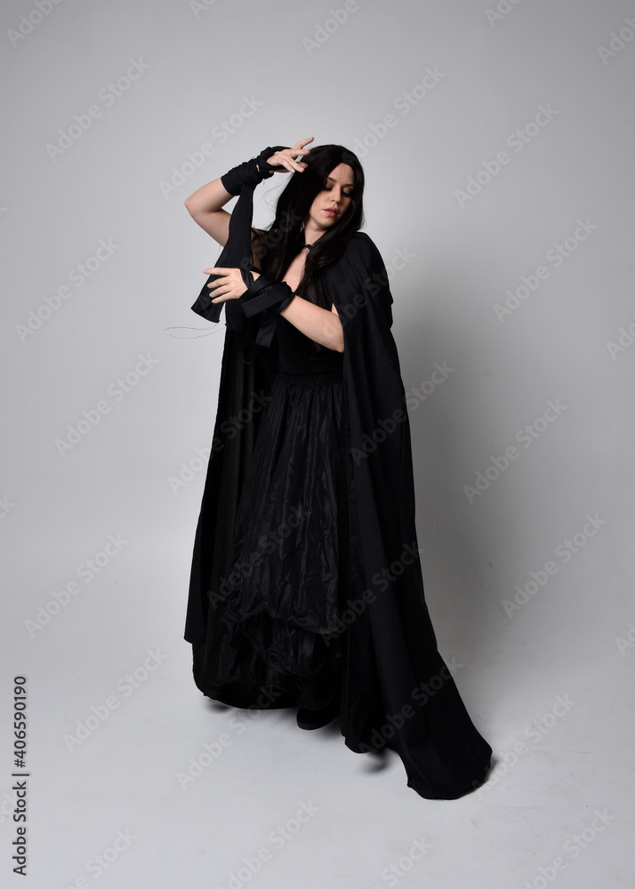 Full length portrait of pretty black haired woman wearing long dark gown and a cloak.  Standing pose facing away from the camera, against a  studio background.