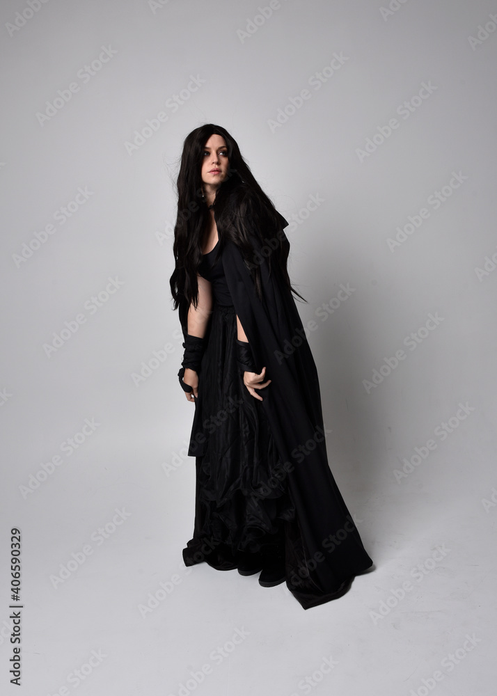 Full length portrait of pretty black haired woman wearing long dark gown and a cloak.  Standing pose facing away from the camera, against a  studio background.