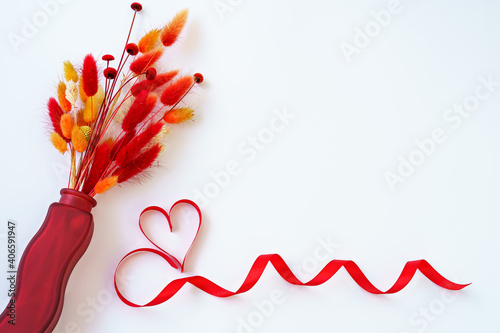 Bouquet of dry red herbs in a vase and a red ribbon in a heart shape on a white background with copy space