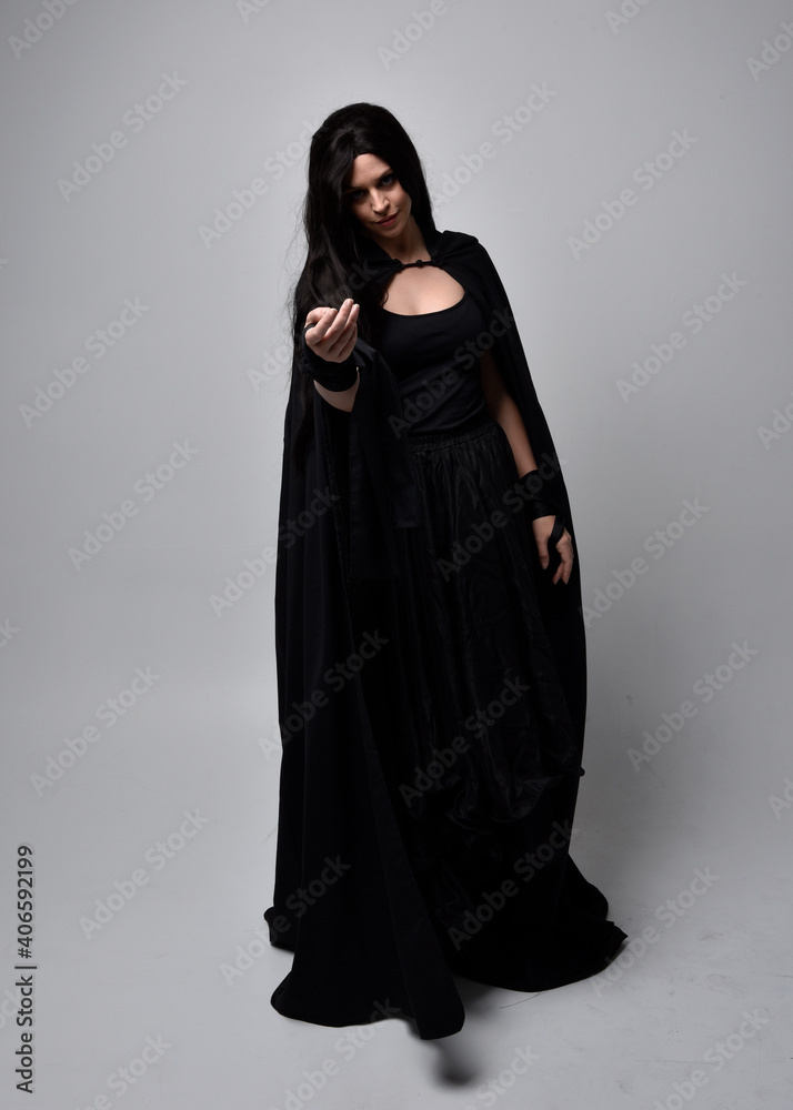 Full length portrait of pretty black haired woman wearing long dark gown and a cloak. Standing pose facing away from the camera, against a studio background. Full length portrait of pretty black haired woman wearing long dark gown and a cloak. Standing pose facing away from the camera, against a studio background.