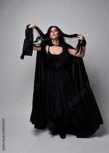 Full length portrait of pretty black haired woman wearing long dark gown and a cloak. Standing pose facing away from the camera, against a studio background.