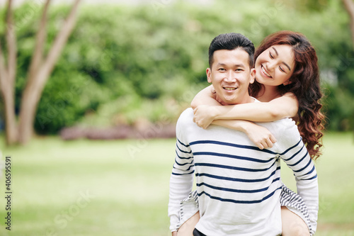 Pretty happy young Asian woman hugging her boyfriend from behind when thay are standing in local park