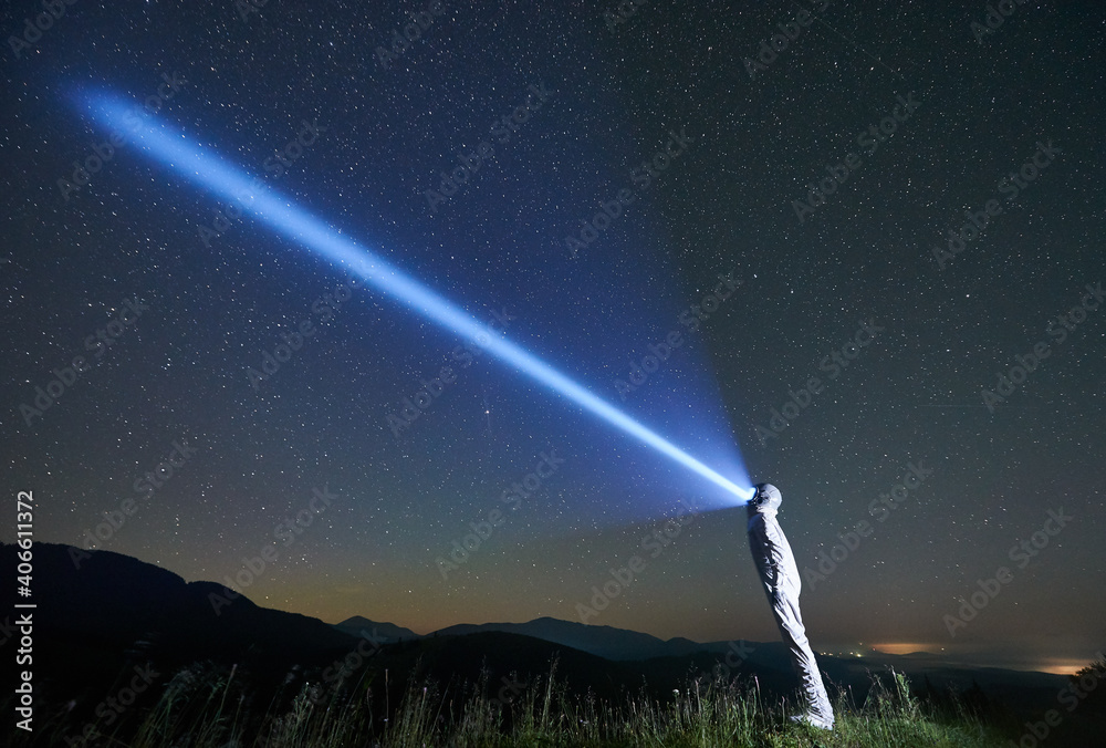 Cosmonaut illuminating fantastic night sky with flashlight. Cosmonaut wearing white space suit and helmet while standing in grassy valley. Concept of light, galaxy and astronautics.