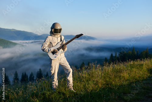 Cosmonaut wearing white space suit and helmet playing white guitar, standing on sunny green mountain glade in summer, morning fog rising up from the valley behind him. Concept of astronautics, music
