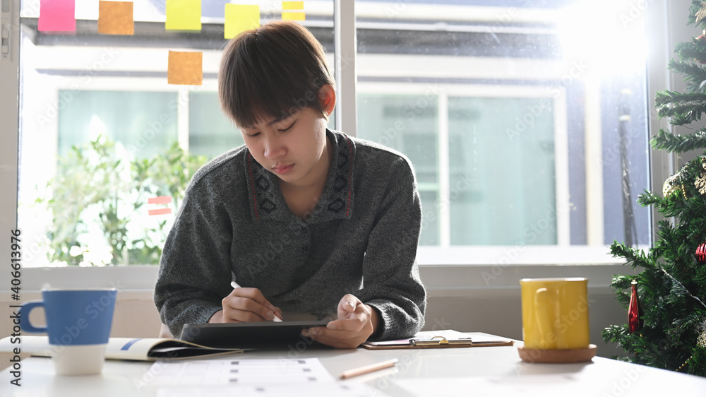 Young female creative designer working on digital tablet at her workspace near window with sun flare. Young female creative designer working on digital tablet at her workspace near window with sun flare.