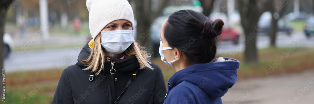 Fototapeta premium Two young women communicating in protective face masks outside. Safety rules during the covid-19 pandemic concept