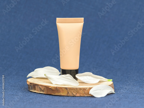 Nude colour blank cosmetic tube with white flower petals and wood cut on blue fabric background. Mock up. Branding concept