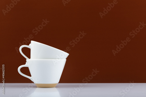 Minimalistic still life with two white coffee cups on in another on white table against brown background with copy space. 