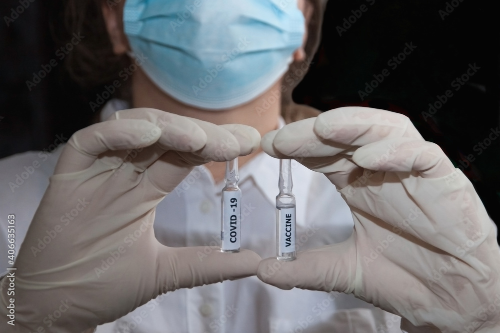 A woman in a white lab coat and medical mask holds two vials of the Covid-19 vaccine A woman in a white lab coat and medical mask holds two vials of the Covid-19 vaccine