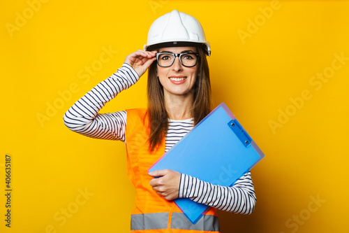 Young woman with a smile in a vest and hard hat holds a clipboard on a yellow background. Concept for construction, new building, renovation. Banner