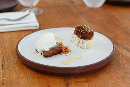 Gourmet dessert, including a honey cake, goat cheese and a honeycomb, served on a flat round stoneware plate with a tall rim. Background of a wood table, set for dinner.
