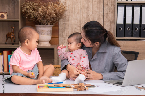 Beautiful Asian single mother happy playing with adorable kids while working at home  children play peekaboo with fun on mother working desk  multi-task young freelancer woman with daughters concept
