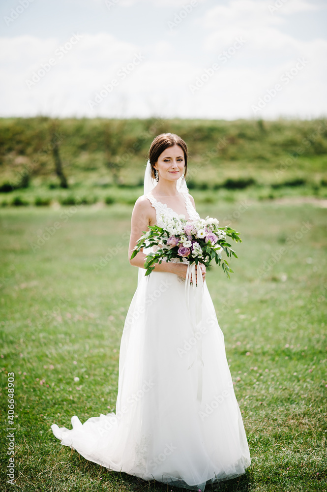 Young beautiful girl in elegant dress is standing and holding hand bouquet of pastel pink flowers and greens with ribbon at nature. The bride holds a wedding bouquet outdoors.