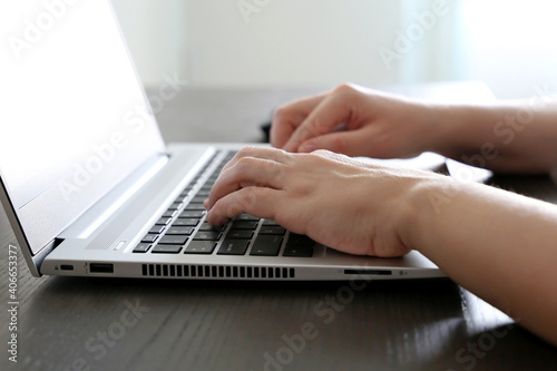 Female hands on laptop keyboard on a desk in sunlight. Woman types on the laptop keyboard sitting near the window, remote work