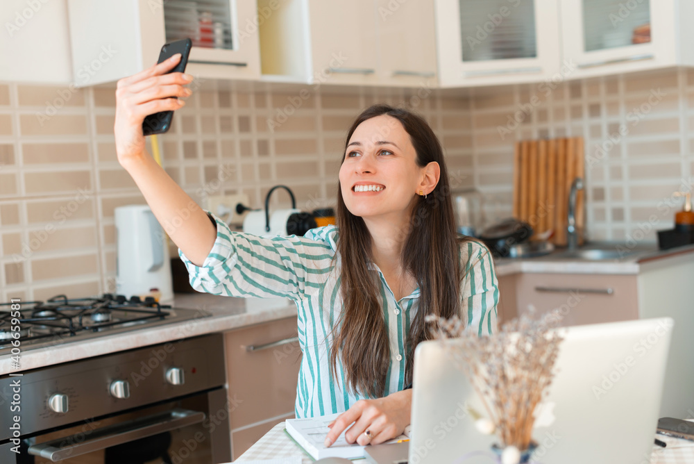 Joyful young woman taking selfie in kitchen while working from home at laptop. Joyful young woman taking selfie in kitchen while working from home at laptop.