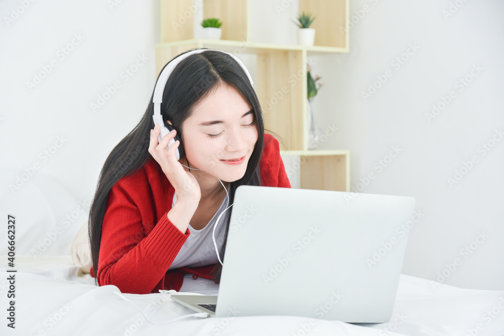 Young asian beautiful girl lying on bed and listen music from notebook computer via headphone.