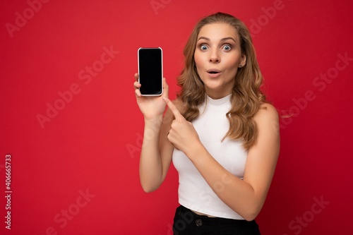 Amazed surprised beautiful young blonde woman wearing white t-shirt isolated on red background with copy space holding smartphone showing phone in hand with empty screen for mockup looking at camera