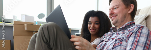 Couple sit on carpet in room and laugh. White man hold laptop on his lap and smile. Black woman sit next to her husband and look at laptop. photo