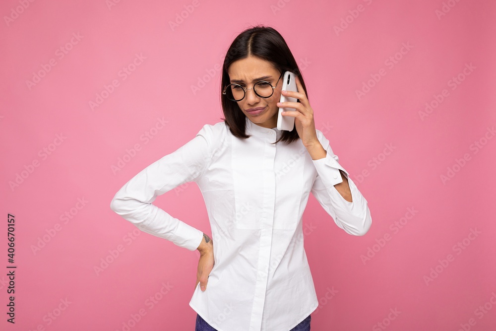 Beautiful dissatisfied young brunette woman wearing white blouse and optical glasses standing isolated over pink background talking on mobile phone looking down Beautiful dissatisfied young brunette woman wearing white blouse and optical glasses standing isolated over pink background talking on mobile phone looking down