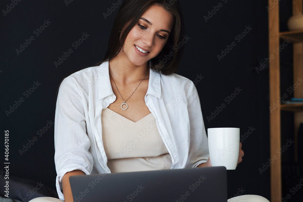 Business woman uses laptop, works remotely from home. Smiling girl in white shirt sits on bed, holding cup of coffee in her hand. Beautiful brunette on dark background of wall in bedroom with laptop. Business woman uses laptop, works remotely from home. Smiling girl in white shirt sits on bed, holding cup of coffee in her hand. Beautiful brunette on dark background of wall in bedroom with laptop.